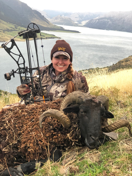 Woman hunting an Arapawa Ram in New Zealand with her bow and arrow and wearing a WILD beanie from River to Ridge Brand
