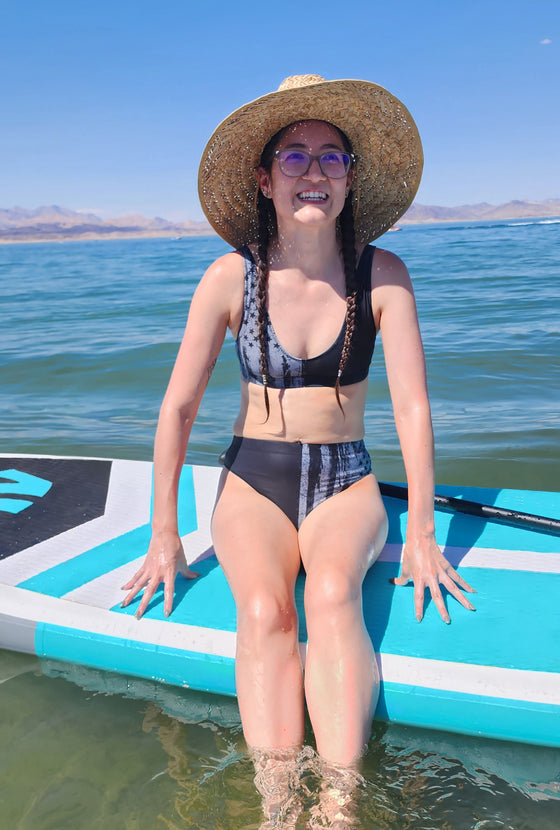 Woman on a paddle board wearing a high waist bikini with a grey flag on it USA, and a straw hat at lake mead in nevada. Swimsuit from river to ridge brand