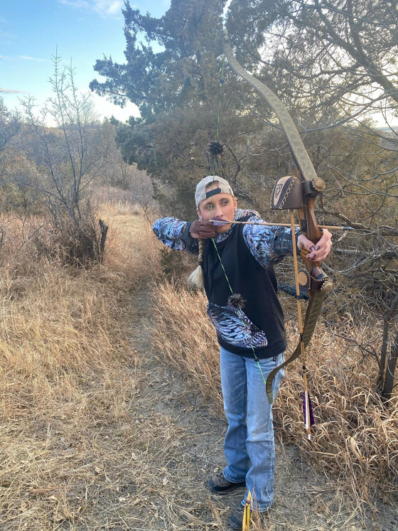 Woman shooting traditional archery with a bow and arrow and wearing a turkey feather hoodie from River to Ridge Clothing Brand