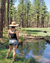 Woman in the forest in northern arizona by a creek wearing a straw hat and turkey feather shorts and a tank top for river to ridge brand