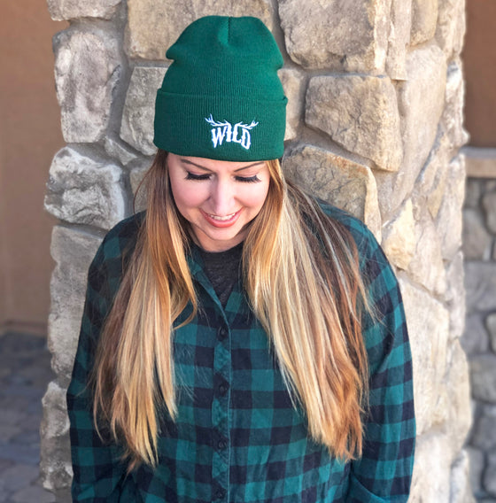 Woman wearing a wild logo beanie with elk antlers and a green and black flanel shirt, she has red hair