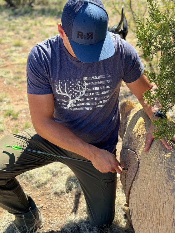 Guy pulling an arrow out of a 3D target wearing a Blue R2R Logo hat and a blue whitetail flag T shirt from River to Ridge Clothing Brand, Archery Compound Bow on the ground
