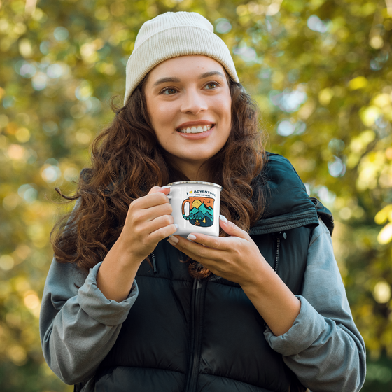 Woman hiking and holding a coffee mug with I love adventure and coffee logo on it. Wearing a beanie in the woods.