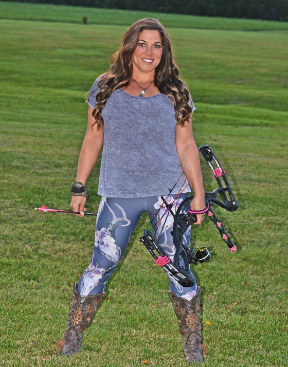 Woman with long brown hair wearing deer antler leggings and holding an archery bow and arrow and wearing cowboy boots and a grey shirt