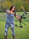 Woman shooting a bow and arrow, archery, wearing deer antler leggings with whitetail skulls and flowers on them and cowboy boots