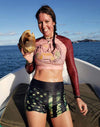 thin woman on a boat on the ocean with a sea shell wearing a blush colored crop top rash guard sun shirt with a tent and cactus and arizona red rocks on it and camo flag shorts