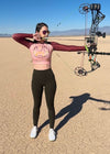 Woman shooting her bow and arrow wearing a crop top rash guard upf shirt from river to ridge brand in blush with a desert camping logo on it and a cactus and tent, photo taken in death valley