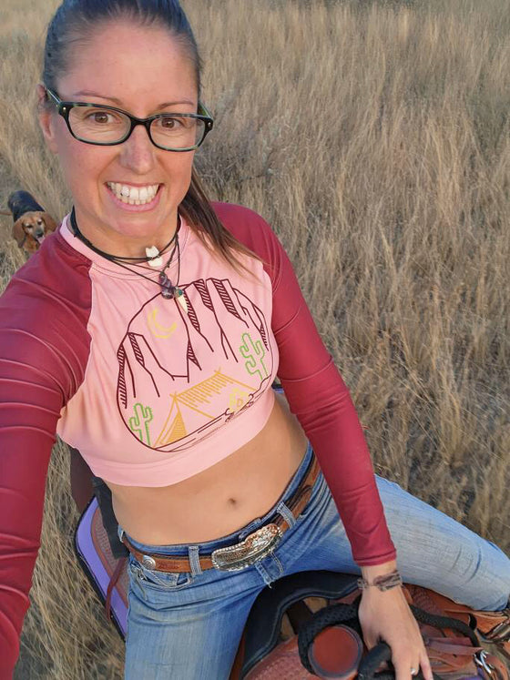 Woman riding a horse with a desert vibes crop top upf sun shirt in peach with a tent and saguaro cactus and sedona red rocks for River to Ridge brand clothing