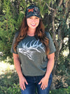 Woman with red hair wearing a green elk logo t shirt and a hunting hat in camo with the word wild on it in hunter orange. she is wearing jeans and standing in the forest