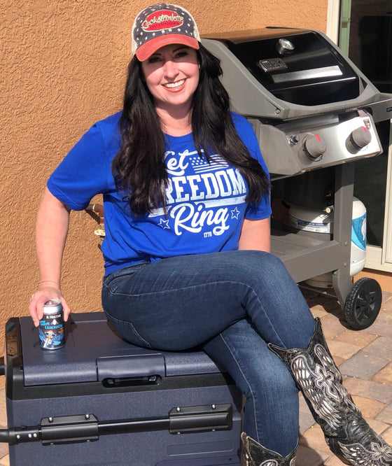 Woman sitting on a cooler with a beer wearing jeans, cowboy boots and a let freedom ring t shirt in blue, with a patriotic flag hat