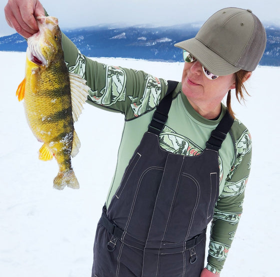 Woman ice fishing catching a perch in idaho in overalls and a trout fishing rash guard shirt in olive