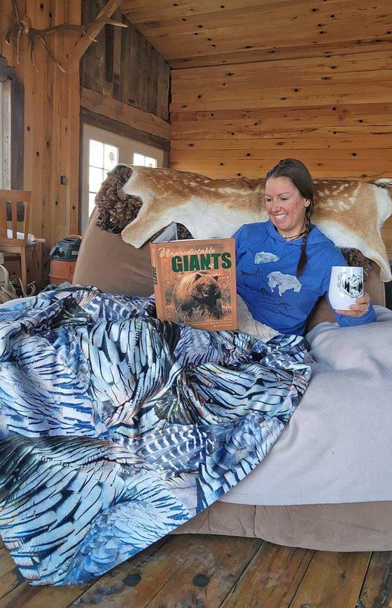 Woman reading a bear hunting book and sitting in a wood lodge with a turkey feather pattern blanket from river to ridge brand