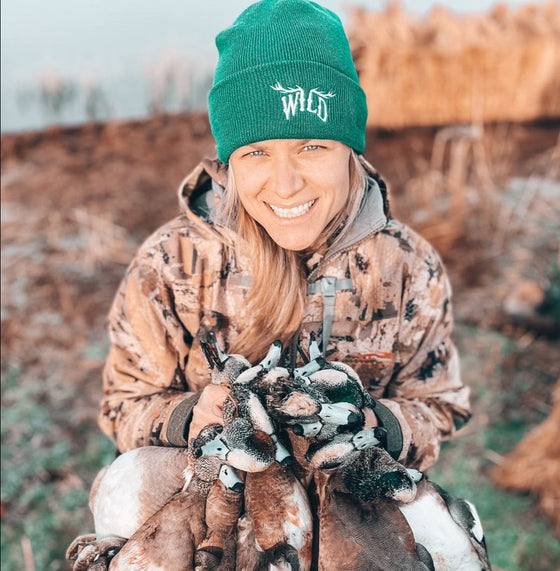 WILD Logo beanie from River to Ridge Brand, featuring the Stay Wild Logo with elk antlers for the 2 center letters. Hat is unisex and is in green and white. Worn by a woman who is duck hunting and wearing camo