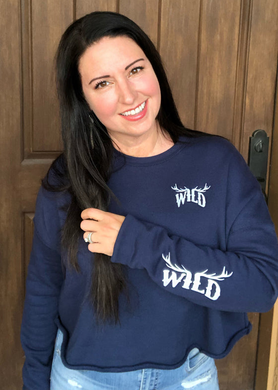 Woman with black hair wearing a cropped blue sweatshirt and jeans. The pullover has a WILD logo on with elk antlers for the letters