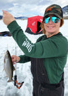 Woman ice fishing on a lake holding up a trout and wearing a WILD sweatshirt in green with an ice fishing hut in the background from the River to Ridge brand