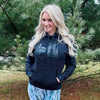 Woman with long blonde hair wearing snow camo leggings and boots and a hoodie in black with a stag deer on it from the Brand River to Ridge - standing in the woods