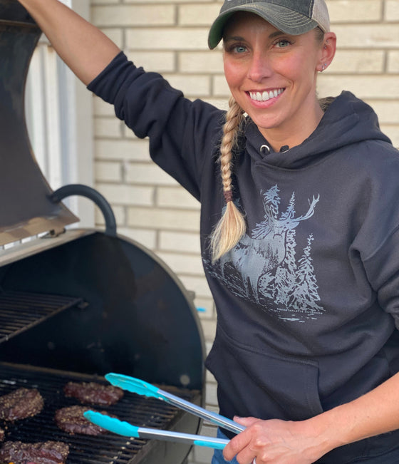 Woman at BBQ cooking burgers on the grill wearing a black woodland logo hoodie with an elk on it from River to Ridge Brand