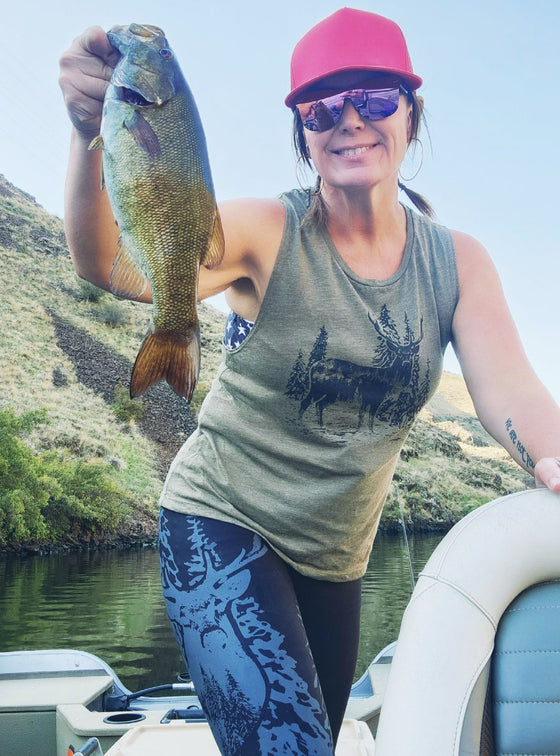 Woman holding up a bass at the lake on a boat wearing a olive muscle tank with a stag on it and also black elk leggings
