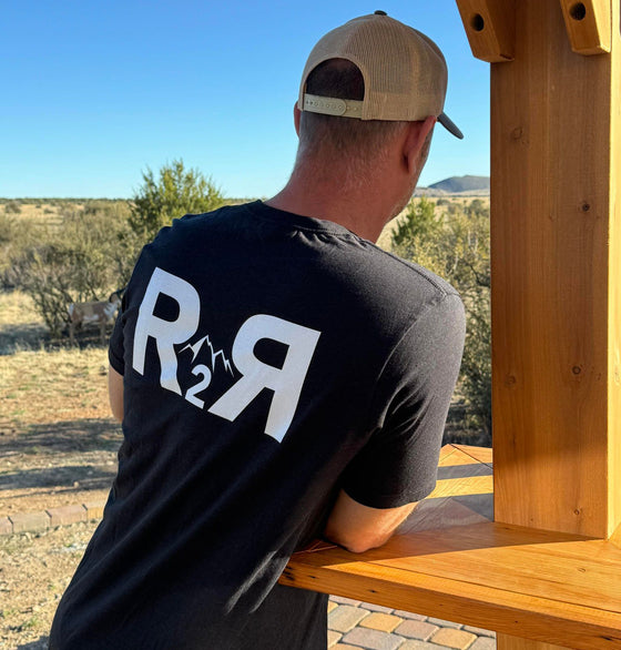 Man leaning on pergola cocktail table outdoors wearing a River to Ridge R2R T shirt in black