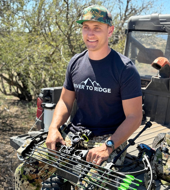 Man wearing a camo flat bill hat from River to Ridge Clothing Brand outdoors sitting on the tailgate of a side by side 4 x 4 offroad and holding his compound bow for hunting