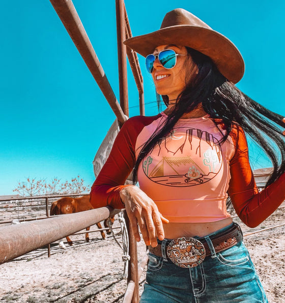 Woman standing at a horse corral wearing a cowboy hat and jeans with rodeo belt buckle and wearing a crop top upf rash guard shirt in maroon and blush pink with a desert vibes logo of a tent and sedona red rocks and a cactus, western shirt for River to Ridge brand