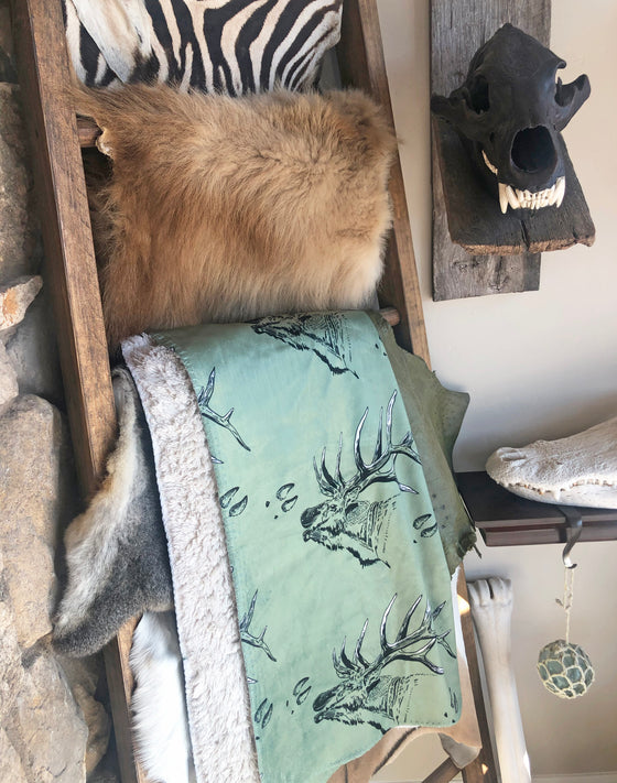 blanket hangin on a blanket ladder with various furs and taxidermy around it and the blanket is olive green with elk antlers printed on it and elk tracks. From River to Ridge brand