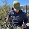 Man working on his compound archery bow wearing a River to Ridge Clothing Brand hat in green and a sportsmans Flag hoodie