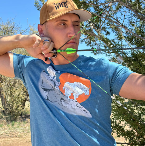 Man shooting archery and wearing a wild logo copper hat and a teal blue mountain goat logo t shirt at full draw. From River to Ridge Clothing Brand