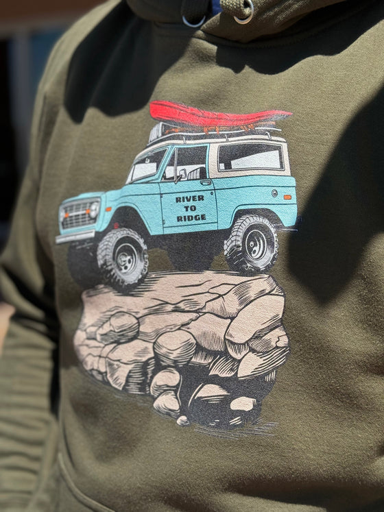 Close up of a guy wearing a River To Ridge Brand hoodie with a vintage classic Bronco on it rock crawling offroad with a red kayak tied on top of it