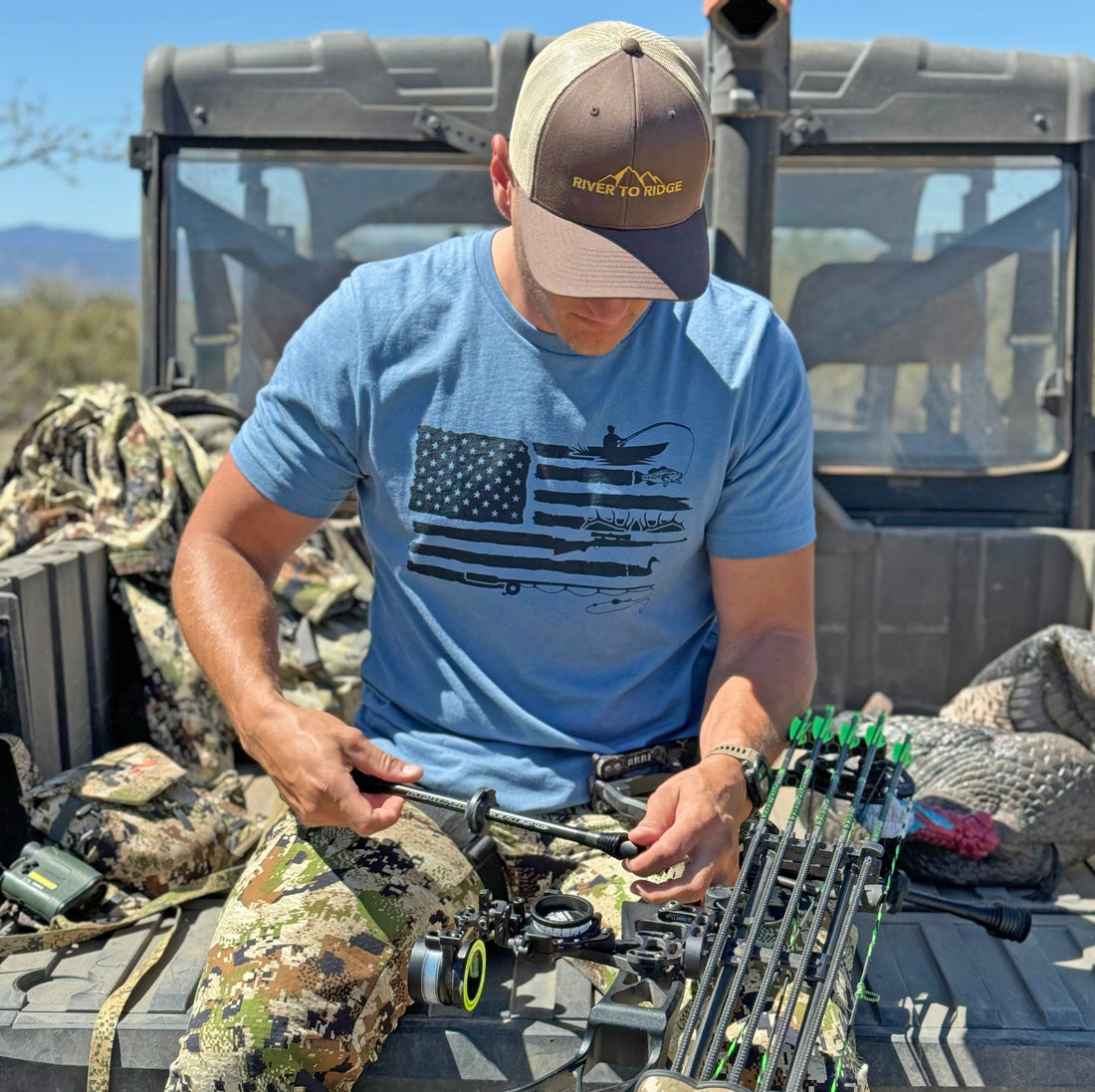 Man wearing a Sportsmans Flag T Shirt from River to Ridge clothing brand in blue and sitting on the tailgate of a 4x4 side by side working on his archery compound bow in camo pants