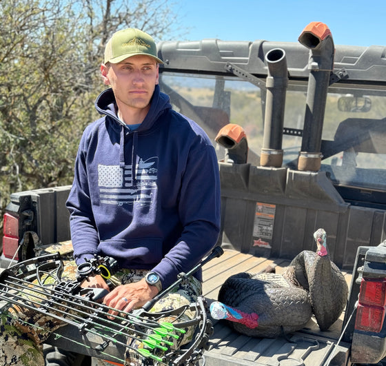 Guy wearing a Sportsmans Flag Hoodie in blue from River to Ridge Clothing Brand while sitting on the tail gate of a side by side 4 wheeler and working on his archery bow. Hoodie has a USA flag on it with outdoor designs added to the stripes like bass fishing, antlers, fly fishing, duck hunting etc..