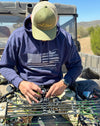 Guy wearing a Sportsmans Flag Hoodie in blue from River to Ridge Clothing Brand while sitting on the tail gate of a side by side 4 wheeler and working on his archery bow. Hoodie has a USA flag on it with outdoor designs added to the stripes like bass fishing, antlers, fly fishing, duck hunting etc..