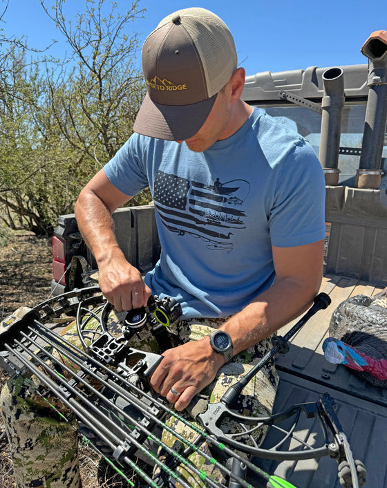 Man wearing a Sportsmans Flag T Shirt from River to Ridge clothing brand in blue and sitting on the tailgate of a 4x4 side by side working on his archery compound bow in camo pants