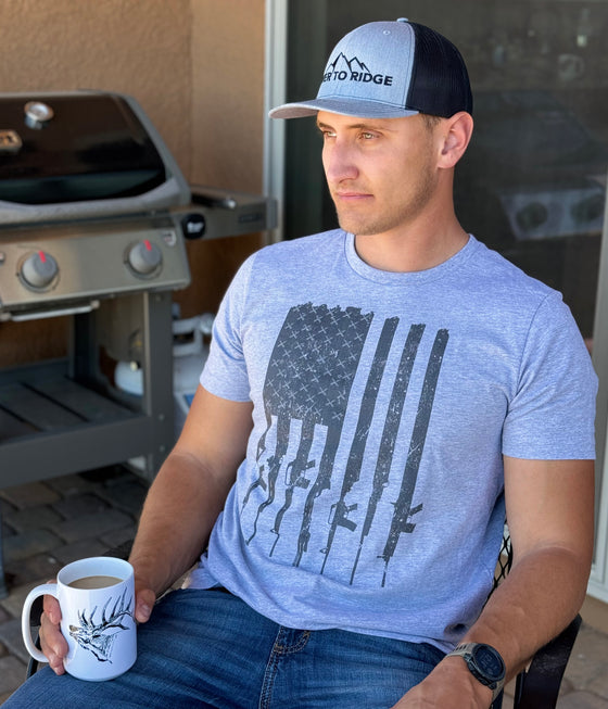 Man wearing a tactical patriotic t shirt in grey sitting and drinking coffee from an Elk Logo coffee mug and wearing a River to Ridge Clothing Brand hat on the patio