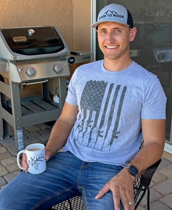 Man wearing a grey and black River to Ridge Logo Hat and a Tactical USA T shirt drinking coffee on the patio from a mug with an elk on it