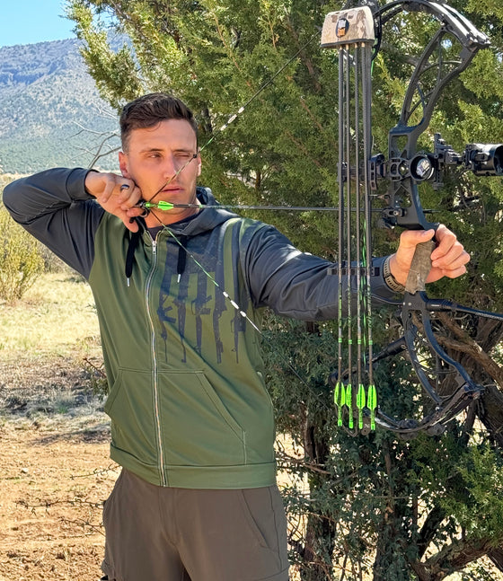 Man shooting a compound archery bow at full draw wearing a tactical logo zip up hoodie in olive and grey with rifles on it from the Brand River to Ridge Clothing