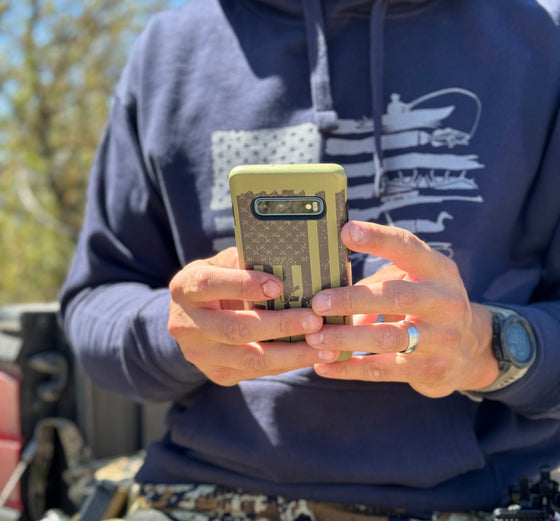Man holding a tactical rifle logo phone case for samsung cell phone, tough case from River to Ridge Brand. Olive and grey, in the background you can see he is wearing a sportsmans flag hoodie in blue