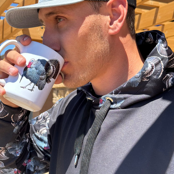 Man drinking from a strutting tom turkey coffee mug from River to Ridge Clothing Brand and wearing a turkey hunting hoodie