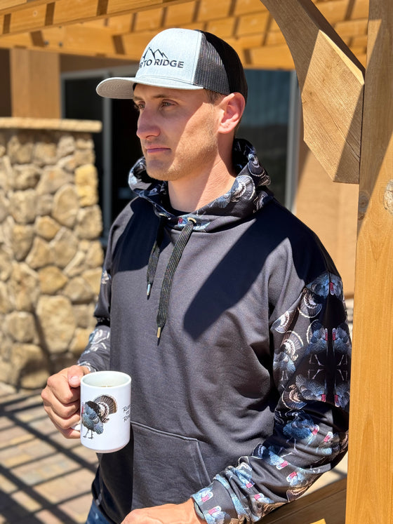 Guy wearing a hoodie that is black with strutting turkeys all over the hood and sleeves in a pattern with a long beard and tail feathers up, he is drinking coffee from a turkey coffee mug and standing outside by a pergola. All from River to Ridge Clothing brand including the grey and black logo hat