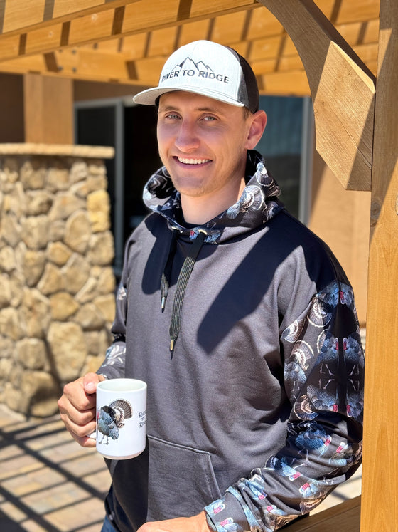 Guy wearing a hoodie that is black with strutting turkeys all over the hood and sleeves in a pattern with a long beard and tail feathers up, he is drinking coffee from a turkey coffee mug and standing outside by a pergola. All from River to Ridge Clothing brand including the grey and black logo hat