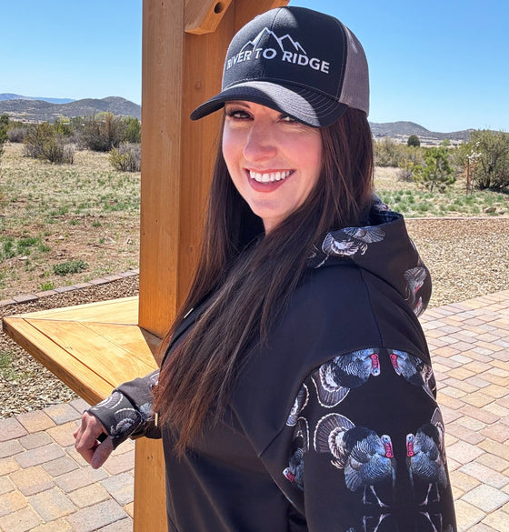 Woman wearing a turkey hunting hoodie and a River to Ridge Logo black and grey snapback trucker hat