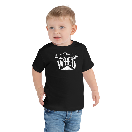 Little boy wearing a toddler t shirt and jeans. The t shirt is from river to ridge brand and is black with a white stay wild logo on it with elk antlers and a mountain
