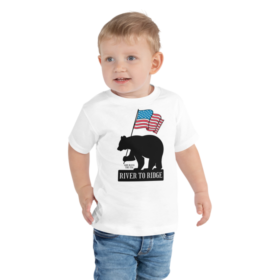 Patriotic Bear Logo from River to Ridge Brand on a toddler t shirt for kids in white. Features a black bear holding a USA flag. Photo of little boy wearing the t shirt