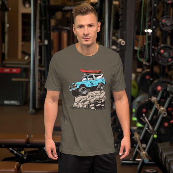 man at the gym wearing a t shirt. Mens offroad classic T shirt in army green, with a graphic of a bronco in blue doing a rock crawl with a kayak on top. From the Brand River to Ridge