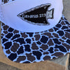 A trucker-style cap with a white front panel and brim, covered in a black and brown camo pattern, resting on a stone surface. The front has a white visor with an embroidered black arrowhead patch containing the text 'GENESIS 27:3'.