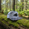 A snapback cap with a brown, black, and white animal print pattern on the front panels and brim, and a solid white mesh back. It features a black leather patch on the front band with the text 'GENESIS 27:3'.