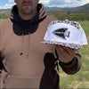 A hand holds a white trucker-style cap with a brown camo brim and back panels in an outdoor grassy landscape under a blue sky. The front patch reads 'GENESIS 27:3' with a archery logo