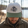 A trucker-style cap with an off-white crown and an olive green brim, featuring a custom patch of a black Labrador retriever holding a duck. The cap is photographed outdoors on a stone surface with a rustic wooden background.