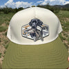 A hand holds a two-tone baseball cap outdoors against a background of grassy terrain, mountains, and a blue sky. The cap features an off-white crown with a black and gray dog graphic on the front, and an olive green brim.
