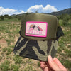 A hand holds a camouflage trucker hat outdoors against a blue sky and grassy landscape. The hat has an olive green front panel and a pink rectangular patch with a bird illustration and the word 'FAITH'.